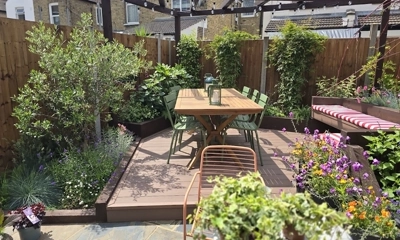 Modern small backyard garden with wooden decking, outdoor dining table, green chairs, striped bench seating, pergola with string lights, and lush plants and flowers – stylish urban outdoor living space