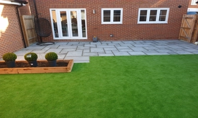 Gray sandstone patio, wood planters embrace lush artificial lawn oasis.
